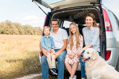 Preview: smiling young family with retriever dog sitting in car trunk and looking at camera in field