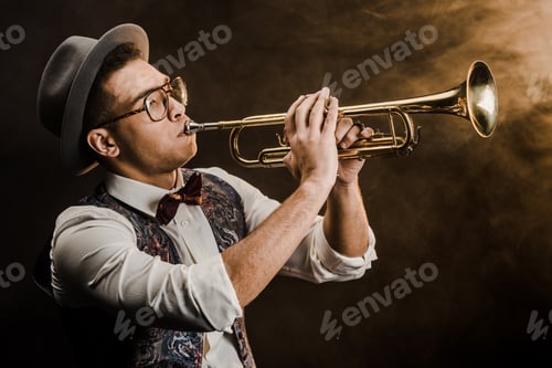 Preview: young mixed race jazzman in hat and eyeglasses playing on trumpet on stage with dramatic lighting