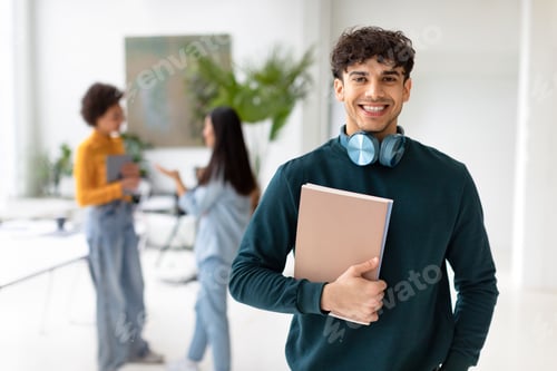 Preview: Happy european student guy standing with workbooks in university classroom and smiling at camera