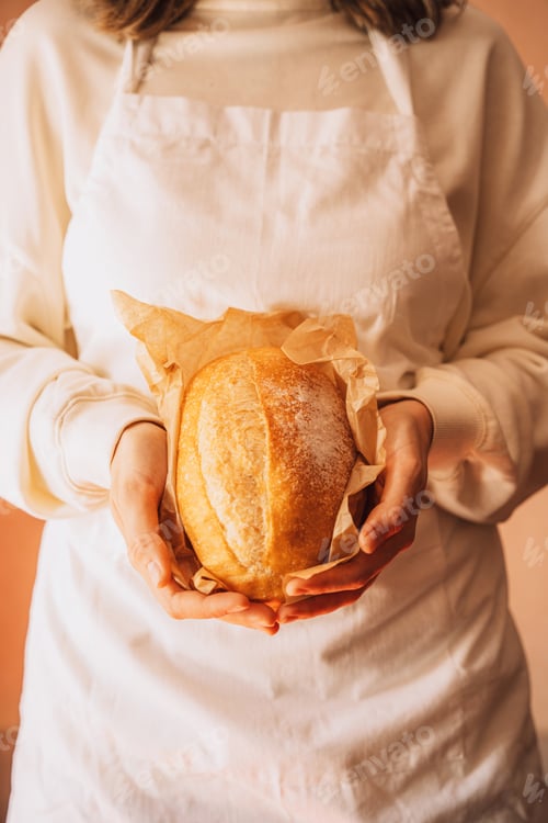 Preview: Fresh loaf of bread in paper packaging in the hands of baker in white apron