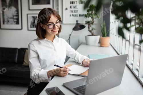 Preview: Happy millennial european businesswoman working on laptop, making notes in cozy coworkign space