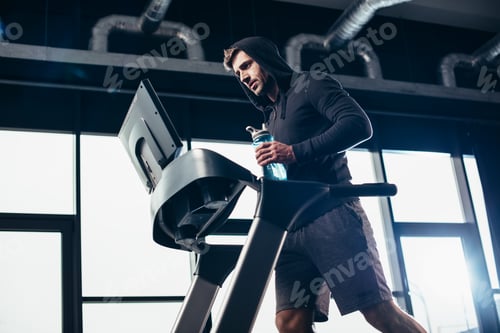Preview: low angle view of handsome sportsman in hoodie exercising on treadmill and holding sport bottle with