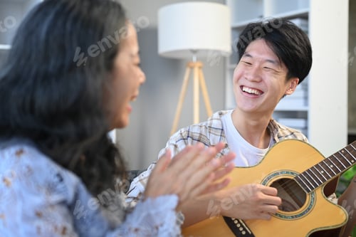 Preview: Young Asian man playing guitar and singing with grandmother at home.