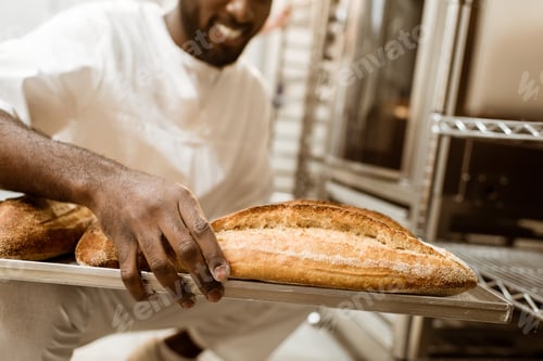 Preview: cropped shot of african american baker with tray of fresh loaves of bread on baking manufacture