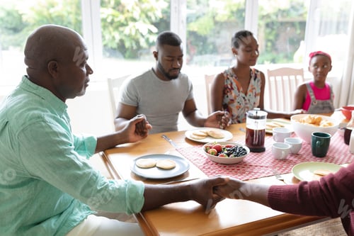 Preview: African american parents, daughter and grandparents sitting holding hands in prayer at dinner table