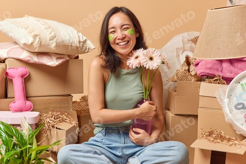 Preview: Positive Asian woman holds vase of gerberas applies green hydrogel patches under eyes to reduce puff