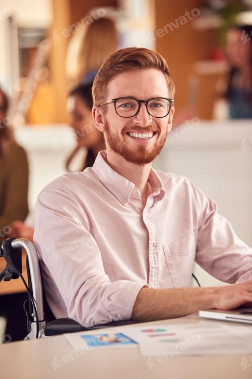 Preview: Portrait Of Businessman In Wheelchair Sitting At Desk Working On Laptop In Busy Office