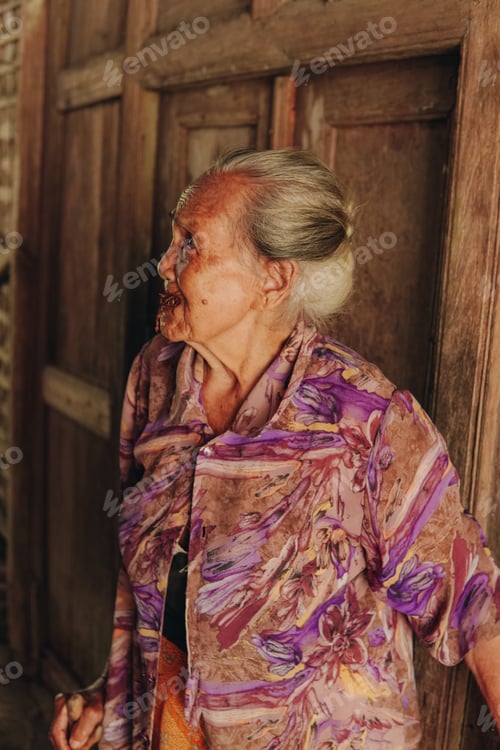 Preview: Senior elderly woman wearing purple kebaya, traditional Indonesian clothes, standing by the door.