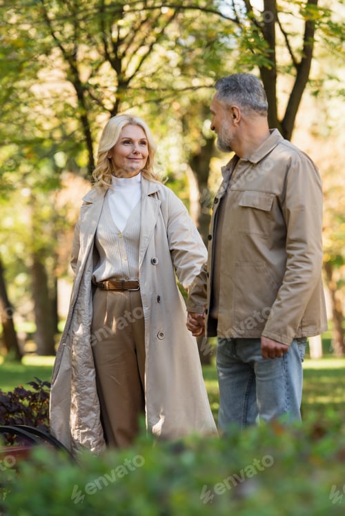 Preview: Positive blonde woman in trench coat holding hand of husband in park