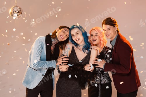 Preview: happy and stylish queer friends embracing and looking at camera under falling confetti on beige
