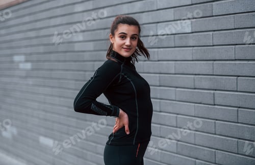 Preview: Young sportive girl in black sportswear standing outdoors near gray wall