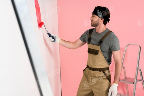 Preview: side view of man in working overall and headband painting wall in red by paint roller near ladder