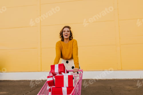 Preview: Satisfied girl posing with shopping cart full of presents. Outdoor photo of woman buying gifts.