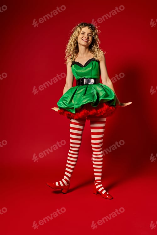 Preview: cheerful blonde new year elf standing still in festive green dress and stockings smiling at camera