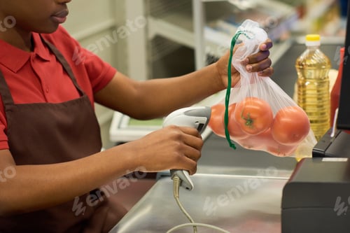 Preview: Young black woman in red shirt and brown apron scanning packed fresh tomatoes