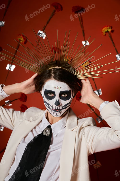 Preview: woman in sugar skull makeup and white suit adjusting festive crown on red backdrop with flowers