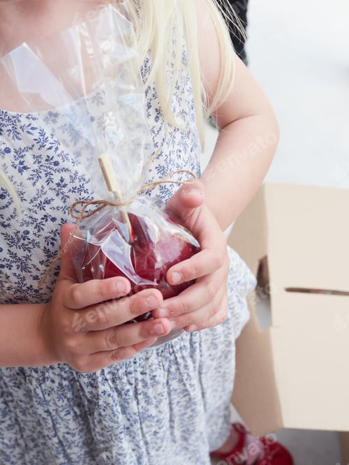 Preview: girl's hands holding a red candy apple wrapped in cellophane paper as a sweet treat