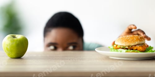 Preview: Overweight black woman looking at hamburger from under table, selecting between healthy and