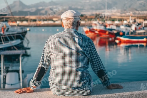 Preview: Back view of relaxed senior man in casual shirt sitting at the sea port at sunset looking the boats
