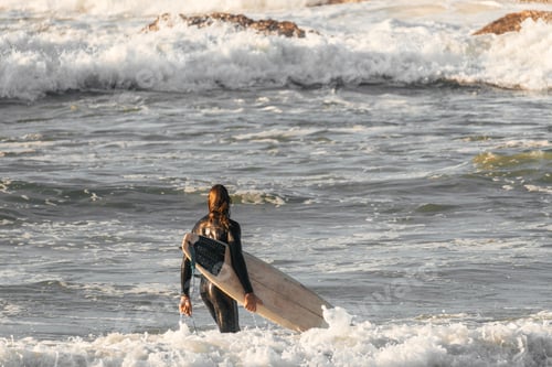 Preview: View from behind of a surfer carrying his surfboard into the water at a time of big waves