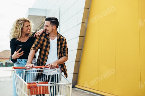Preview: Couple using smartphone while holding a shopping cart while in front of the supermarket or the mall