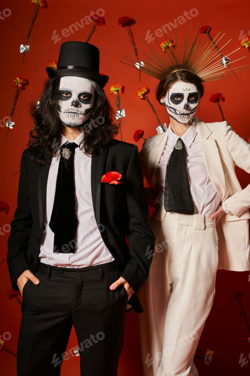 Preview: couple in dia de los muertos makeup and festive outfit posing on red backdrop with carnations