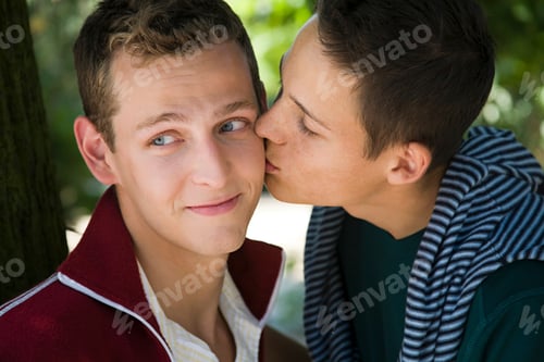 Preview: A tender moment as one teenager kisses another on the cheek under the shade of a tree.