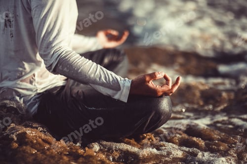 Preview: Candid authentic male millennial sitting in lotus pose gyan mudra in ocean waves in bright sun