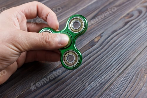 Preview: A man's hand holding and playing metal green color fidget spinner with wooden table background