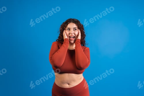 Preview: Excited curvy model showing excitement with open mouth and hands on cheeks on blue background
