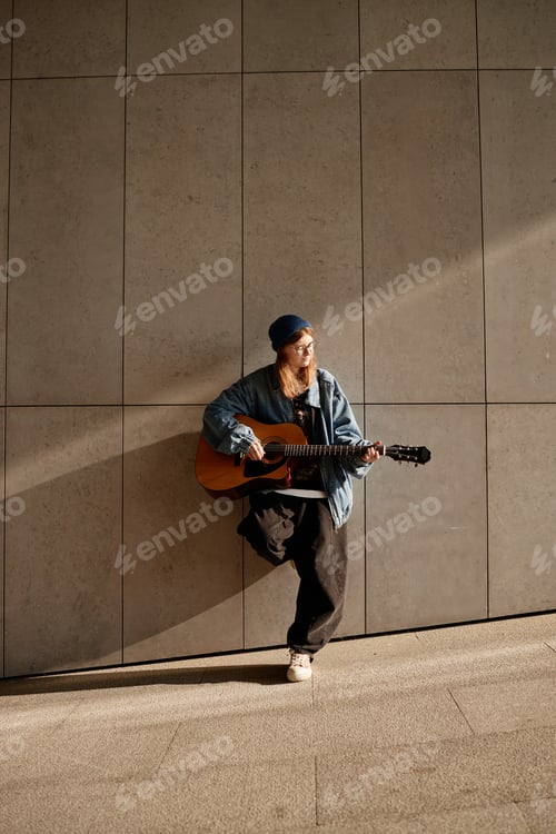 Preview: Young Adult Caucasian Woman Playing Guitar While Leaning Against Wall Outdoors