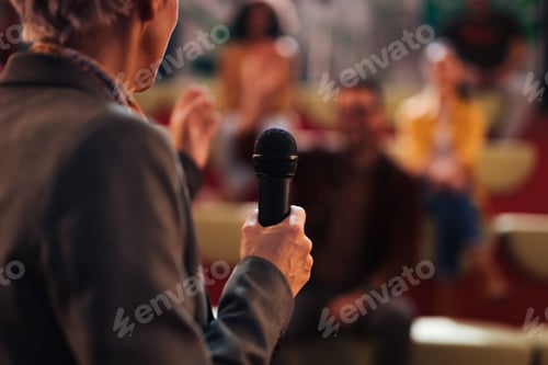 Preview: Focus on the female hand, holding a microphone, people sitting in the background.