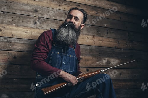 Preview: Vintage Farmer Holding Rifle Standing Against Wooden Wall In Barn.
