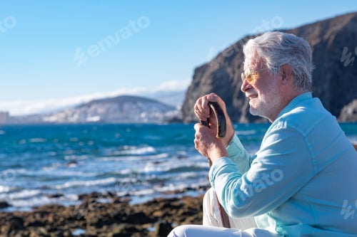 Preview: Senior smiling bearded man sitting at sunset light on a rocky beach face the ocean