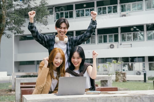 Preview: Three cheerful asian university students are raising their arms in excitement while looking at a lap
