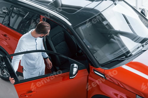 Preview: Young elegant man in white formal clothes standing near red automobile