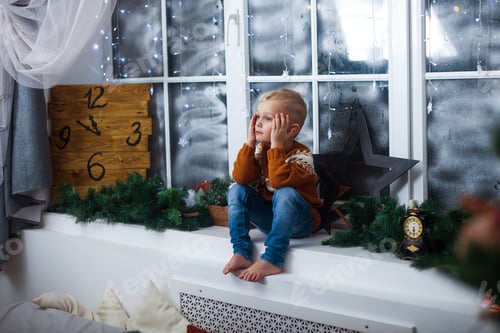 Visualização: menino de suéter está sentado no parapeito da janela de casa, esperando por milagres, presentes e Natal