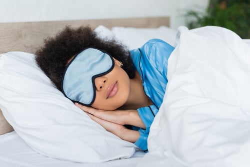 mujer afroamericana rizada en pijama azul y máscara de seda durmiendo en la cama