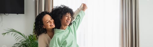 Preview: cheerful african american mother and daughter holding hands while smiling at home, banner