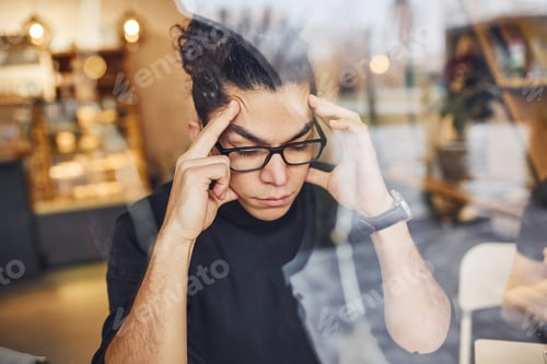 Preview: Young guy with black curly hair sitting indoors in cafe. View through the glass