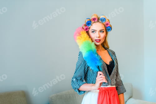 Preview: beautiful blonde smiling housewife in curlers cleaning house with duster