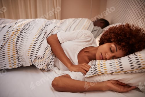 Preview: Millennial African American woman lying on her side asleep in bed, her partner in the background
