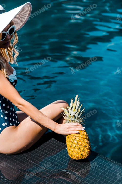 Preview: young woman in swimwear posing with pineapple near swimming pool