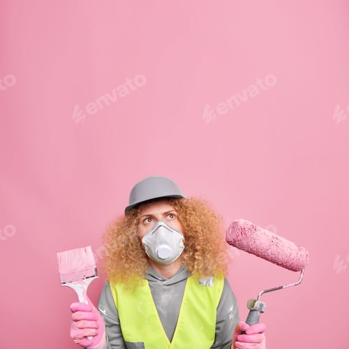 Preview: Serious female builder wears protective helmet and respirator holds painting roller and brush focuse