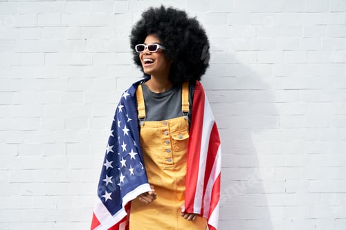 Preview: Happy young African woman wrapped in USA flag standing against white wall.