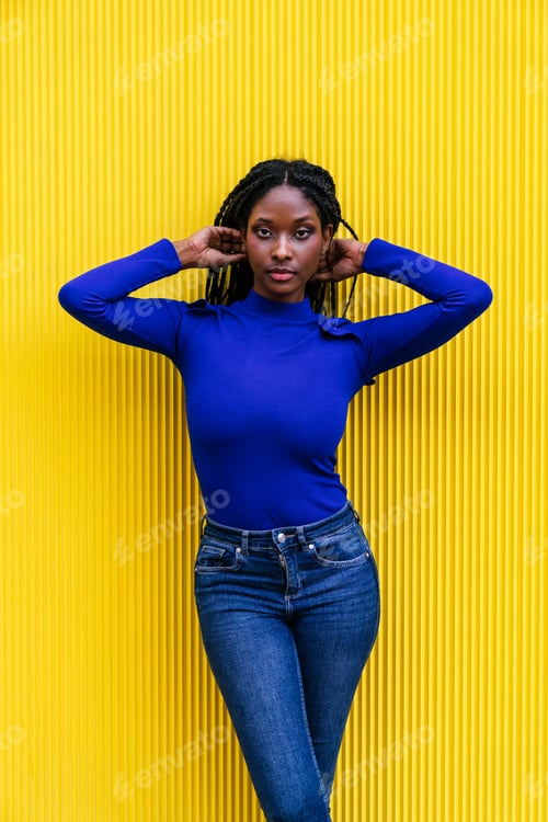 Preview: Afro-American Woman Posing on Yellow Wall, Touching Hair, Wearing Blue Outfit - Vibrant Portrait