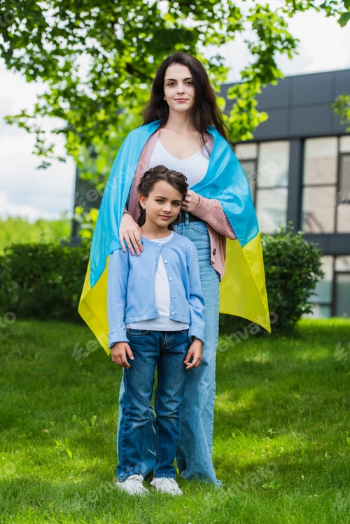 Preview: woman covered with ukrainian flag looking at camera near daughter in park