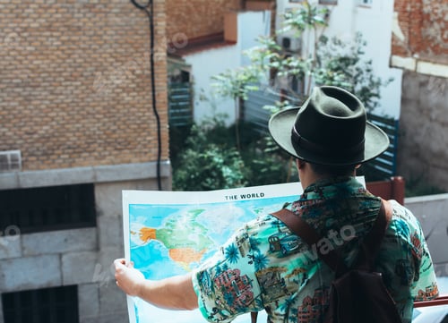 Preview: Bohemian man with mask on his face and hat looking at a map of the world on his balcony.