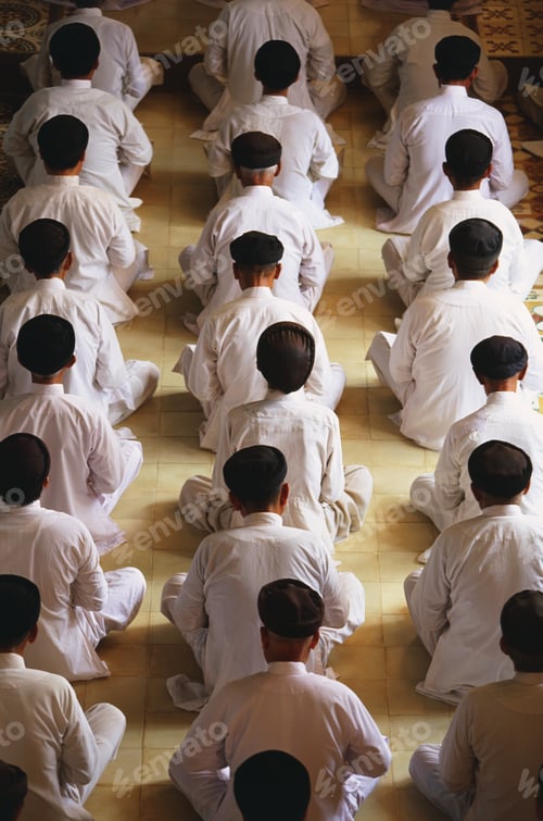 Preview: High angle rear view of of men wearing white robes sitting in rows on floor, praying.