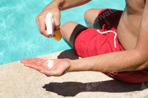 Preview: Unrecognizable young man applying sunscreen sitting at the edge of the swimming pool